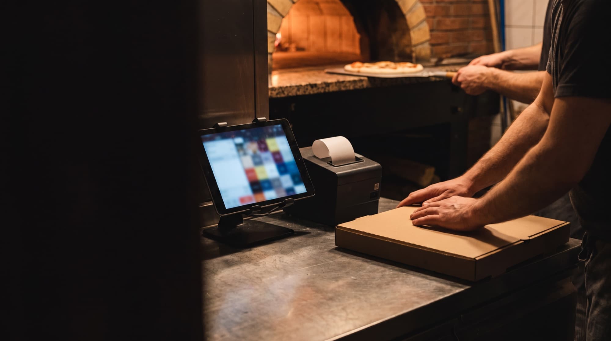 Abendbetrieb hinter der Theke einer Pizzeria: ein Tablet mit Bestellbildschirm, ein kleiner Bondrucker, Hände schließen einen Pizzakarton, dahinter der Steinofen.